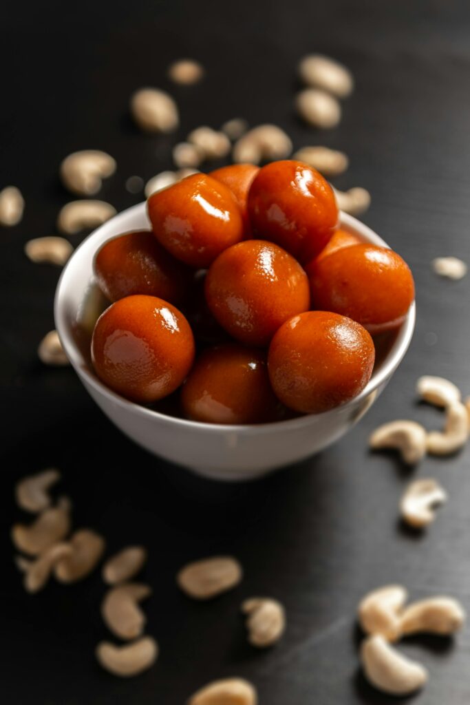 Close-up of fresh gulab jamun in a white bowl surrounded by scattered cashew nuts on a dark background.